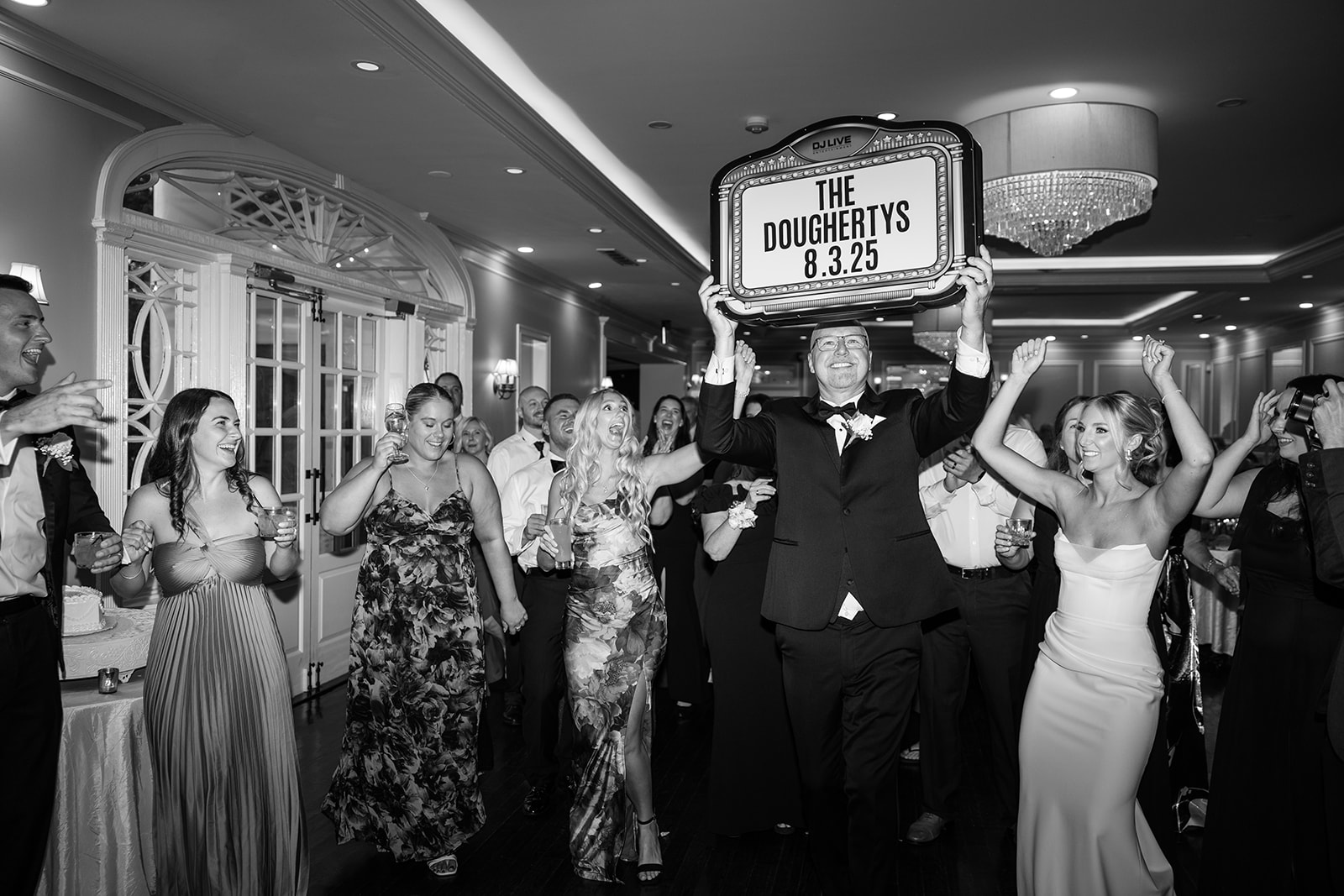 Black and white photo of a guest raising a custom wedding sign on the high-energy reception dance floor.